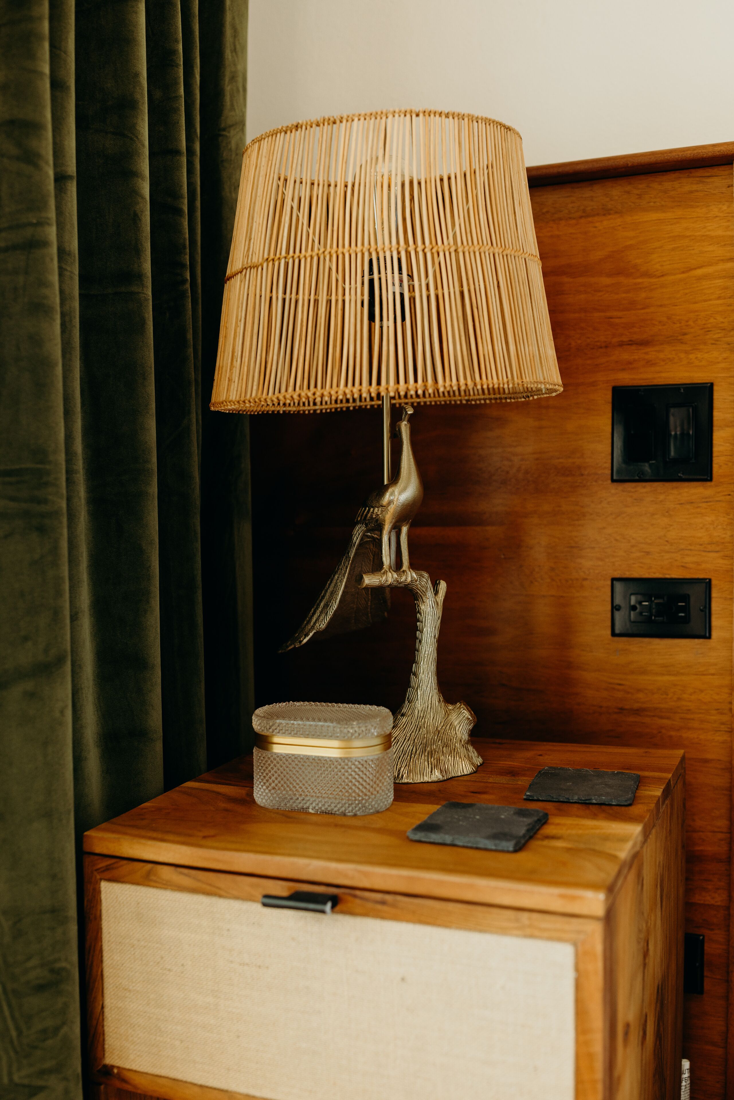 A wooden nightstand with a woven lamp, a small woven basket, and two slate coasters.