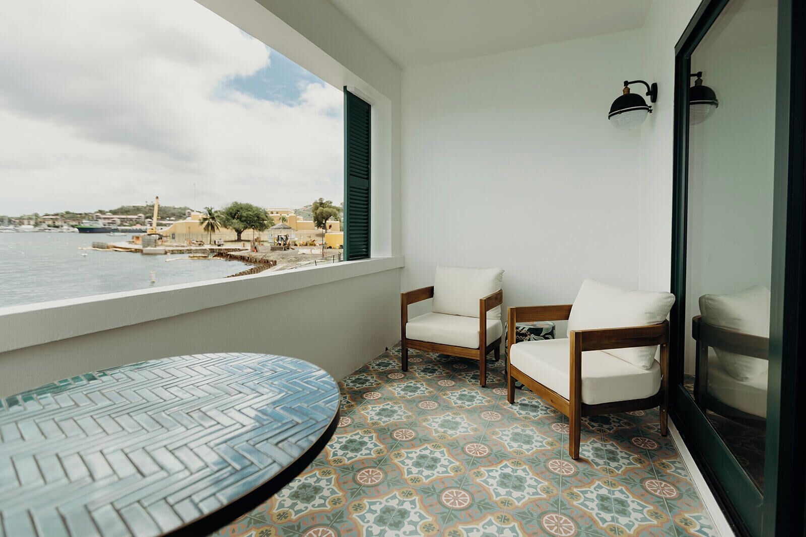 A balcony with two wooden chairs, a round table, and a view of the sea and buildings.