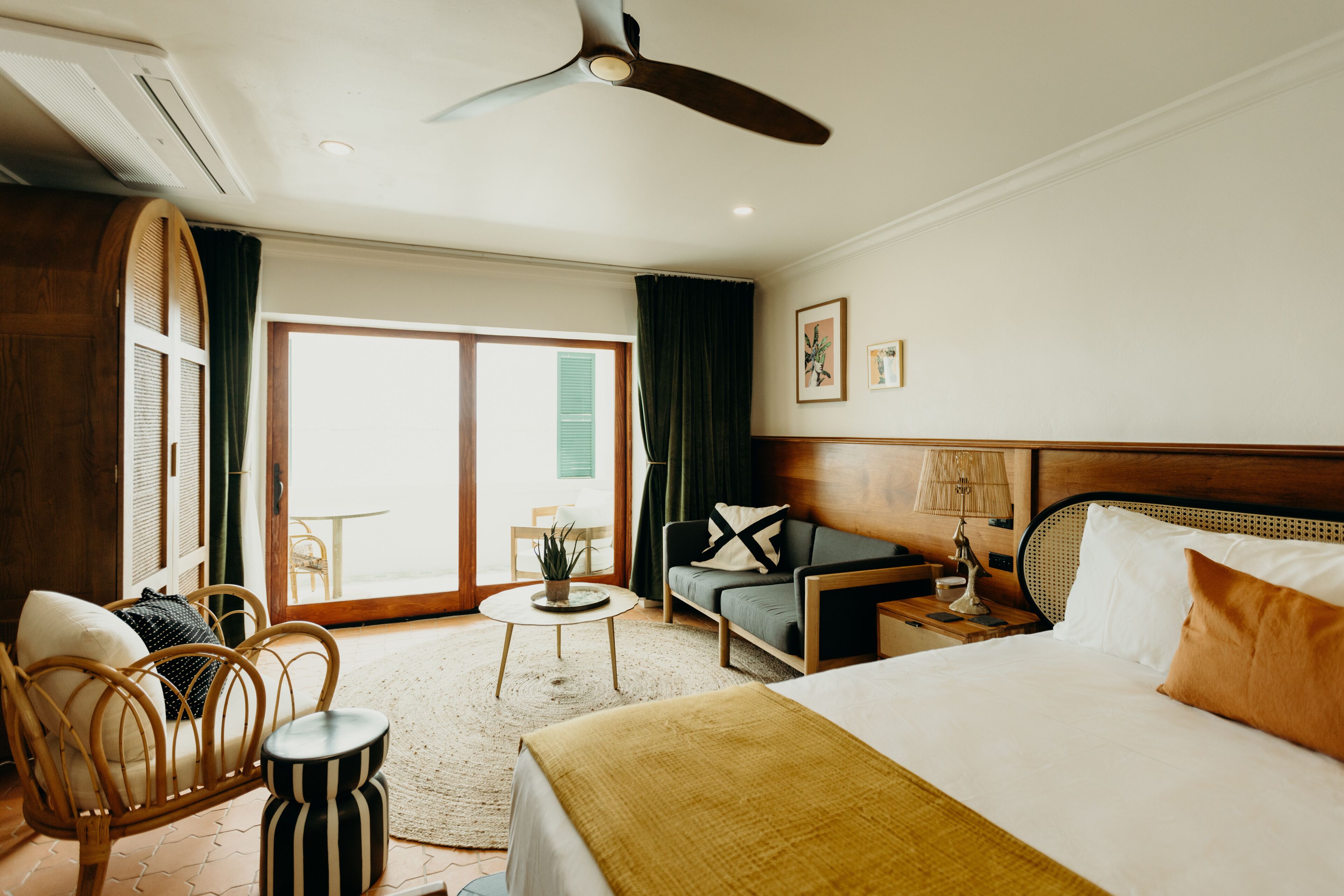 A hotel room with a bed, a chair, a small table, a ceiling fan, and a view of the outdoors.