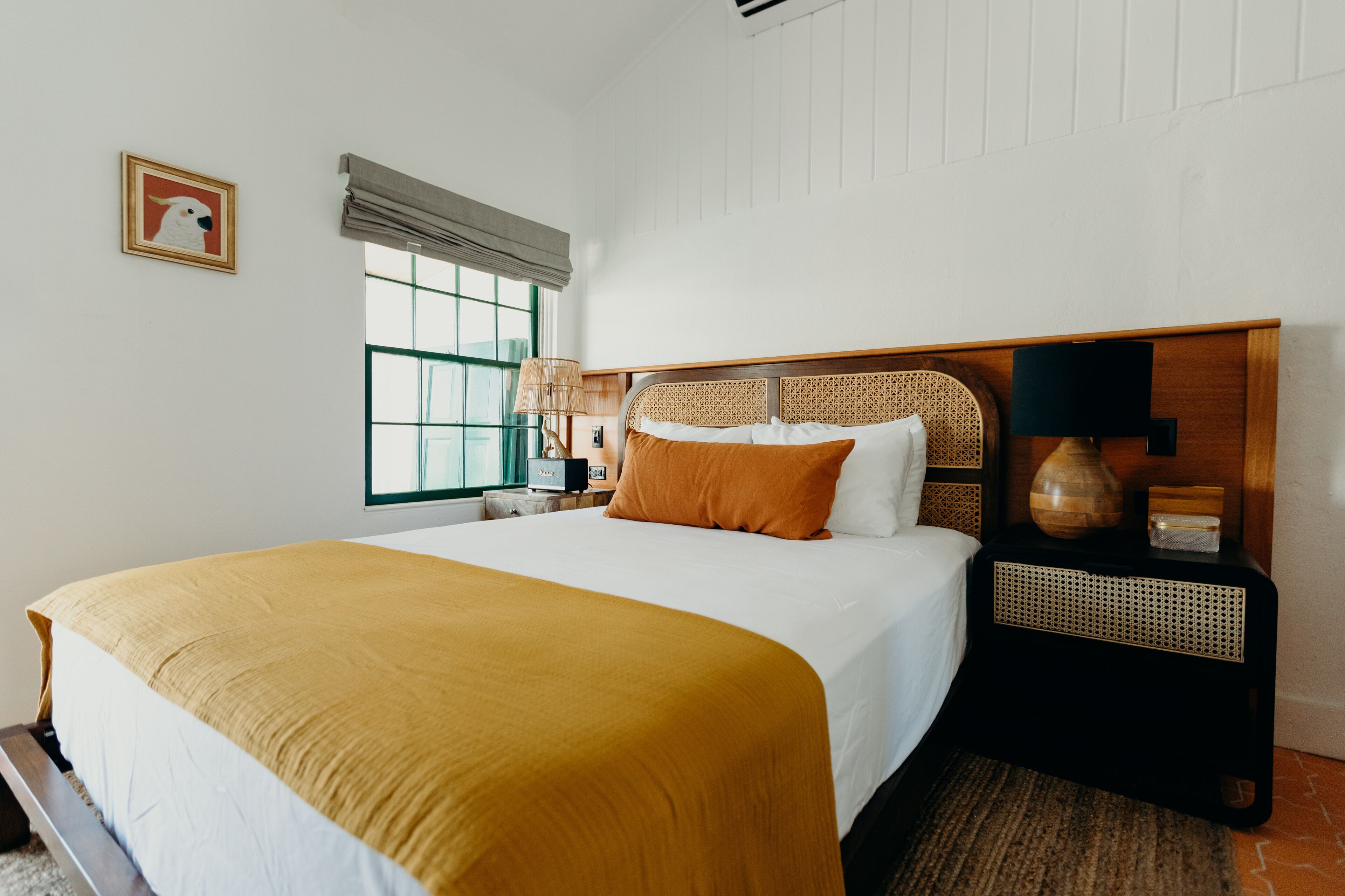 A neatly made bed with a mustard-colored blanket, a wooden headboard, and a bedside table with a lamp and a vase.