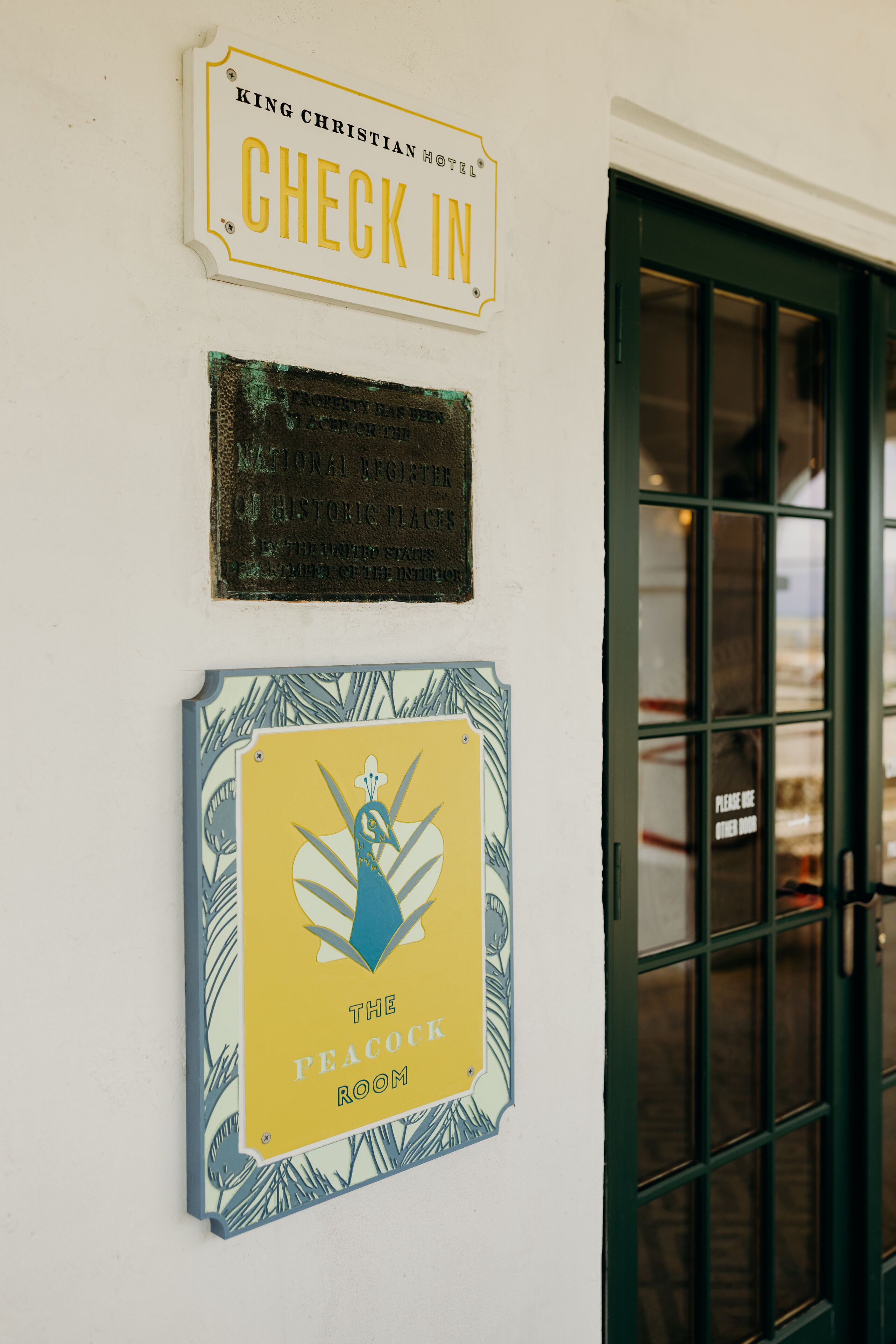 Een muur met drie borden: een 'Check In' bord, een historisch plaquette en een bord met 'The Peacock Room'.