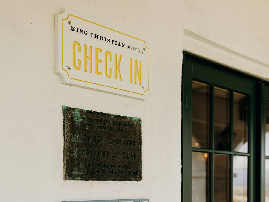 A wall with three signs: a 'Check In' sign, a historical plaque, and a 'The Peacock Room' sign.