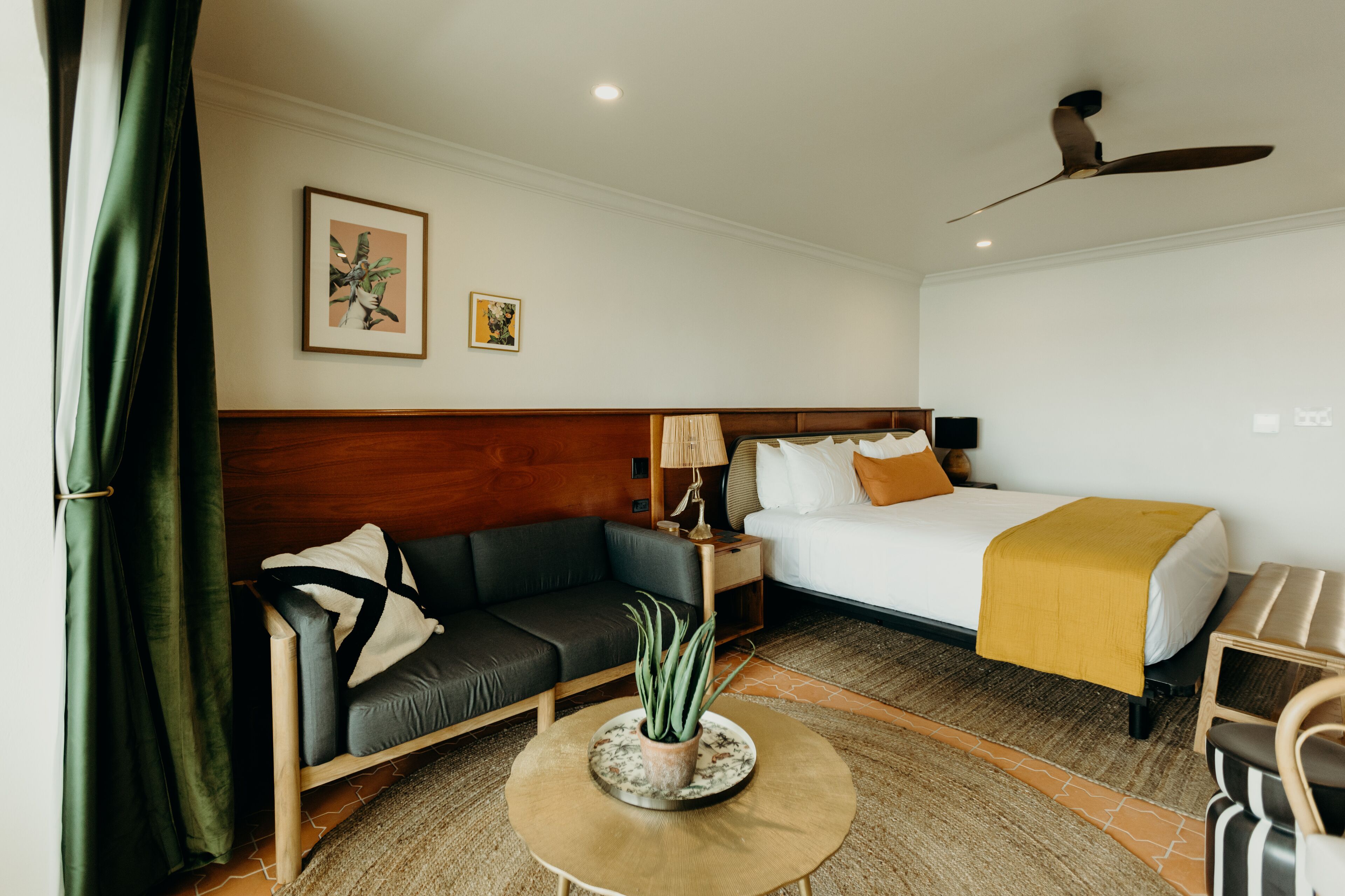 A hotel room with a bed, a sofa, a table with a plant, a ceiling fan, and artwork on the wall.