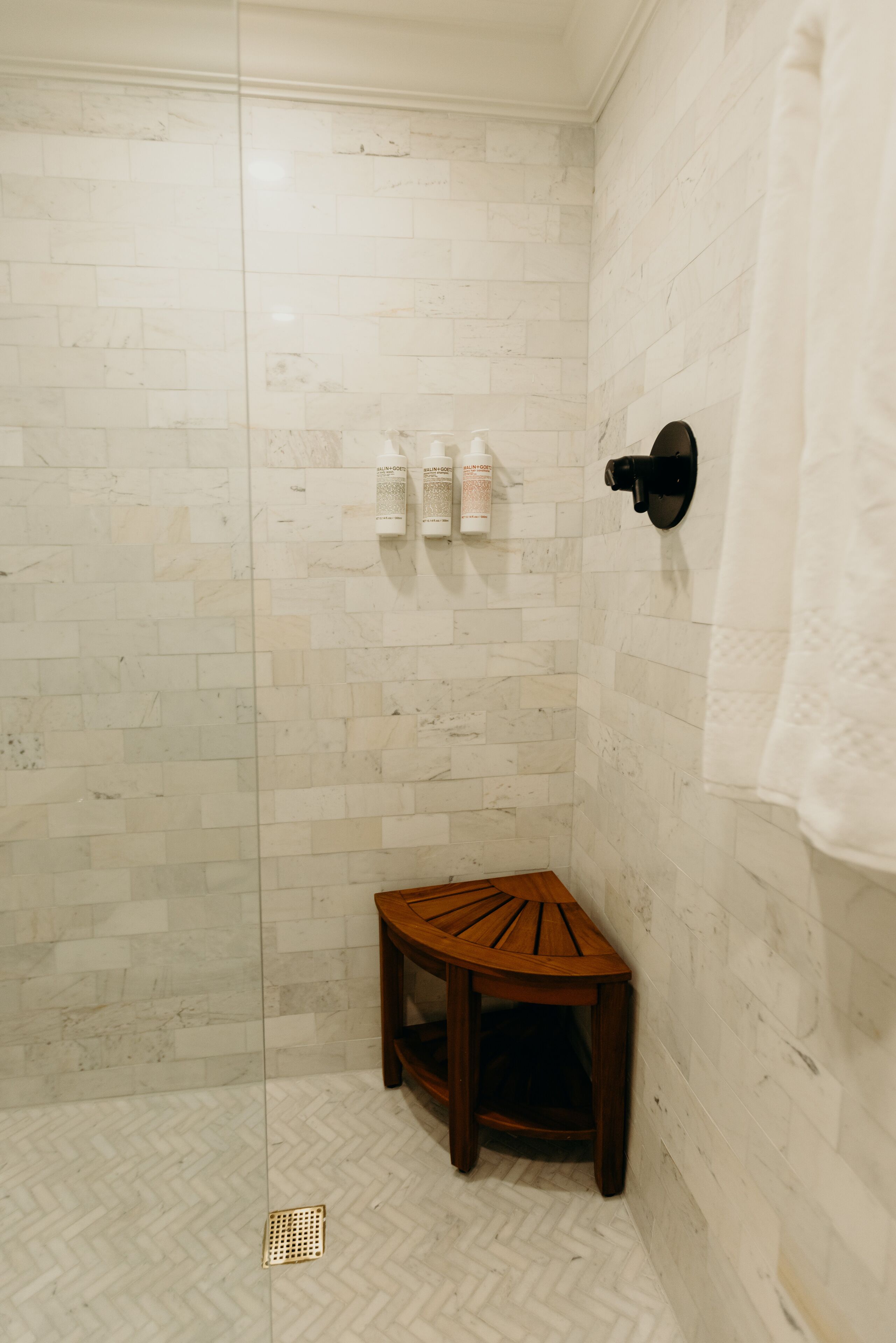 A shower with white marble tiles, a wooden stool, and a towel rack.