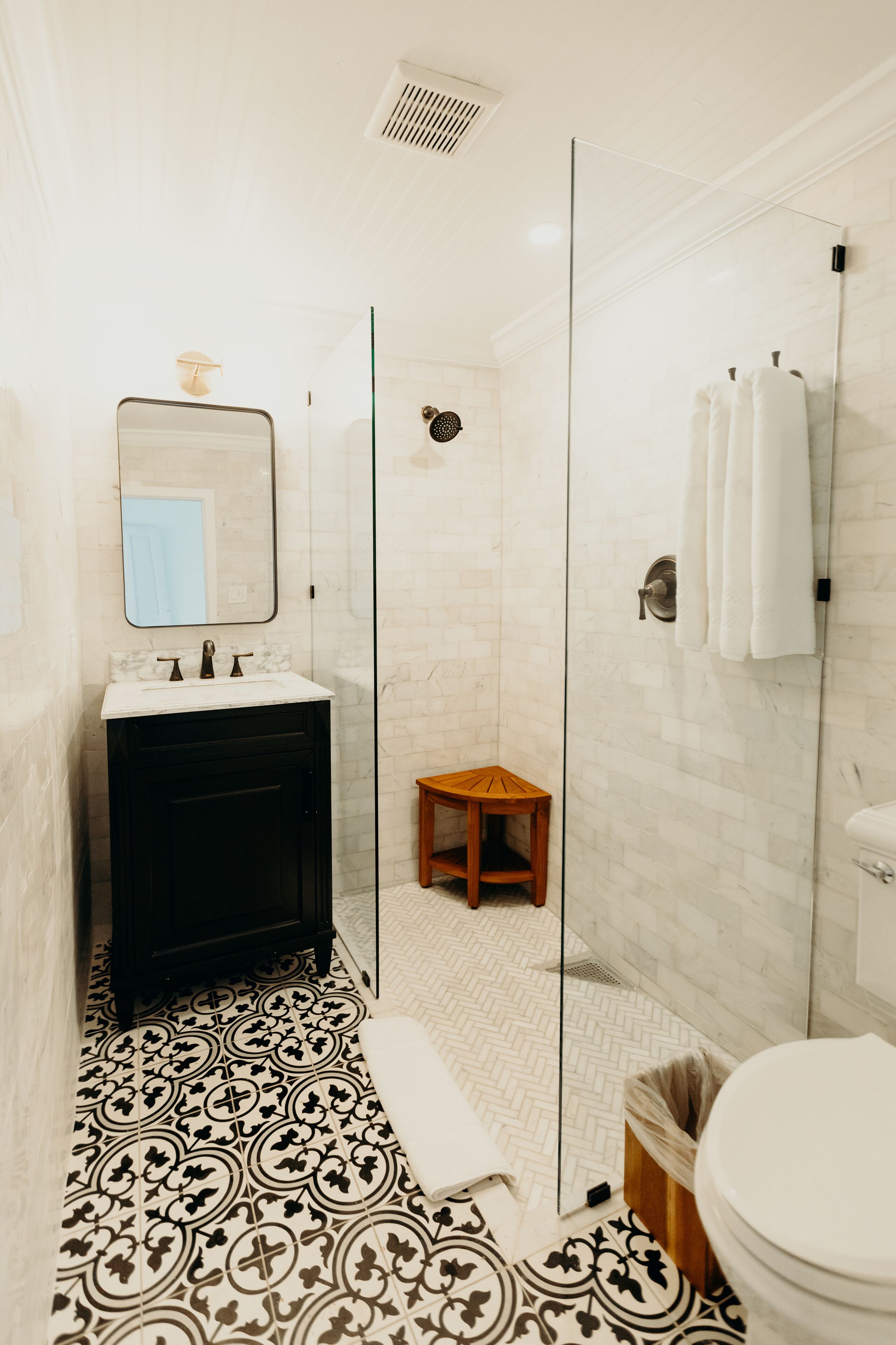 Une salle de bain avec un sol carrelé de carreaux blancs, un meuble-lavabo noir, un miroir, un espace douche et des toilettes.