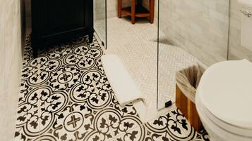 Une salle de bain avec un sol carrelé de carreaux blancs, un meuble-lavabo noir, un miroir, un espace douche et des toilettes.