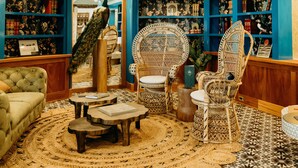 Un acogedor salón de lectura con una amplia estantería azul, dos sillones de mimbre, un sofá capitonado y una alfombra estampada.