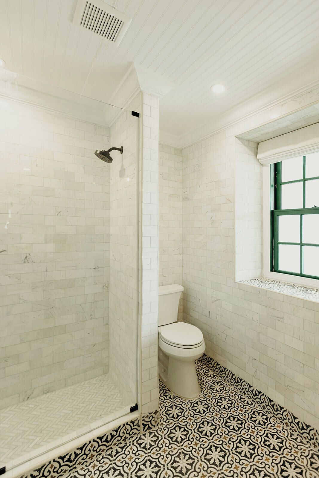 Une salle de bain avec des carreaux de céramique blancs rectangulaires, une douche en verre, des toilettes et un carrelage au motif décoratif.