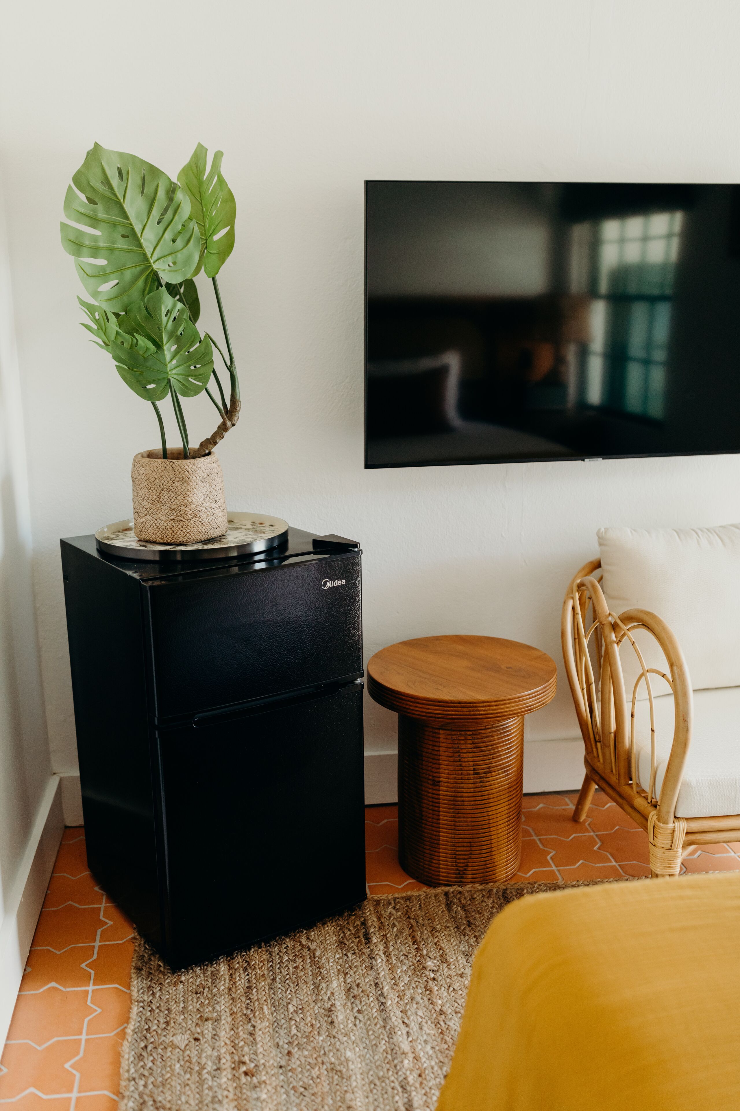 A modern living room with a black speaker, a round wooden stool, a wicker chair, and a large plant on a black stand.