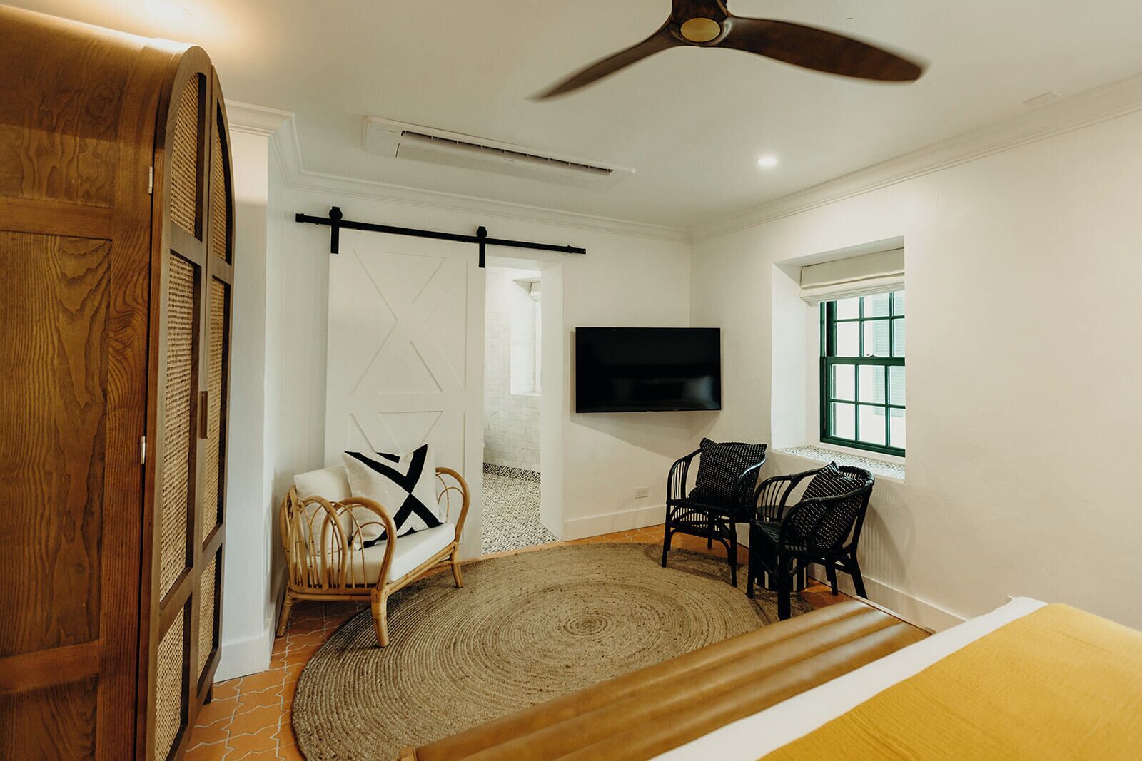 Una habitación con ventilador de techo, televisión montada en la pared, zona de comedor con sillas y mesa, y una puerta grande de madera.