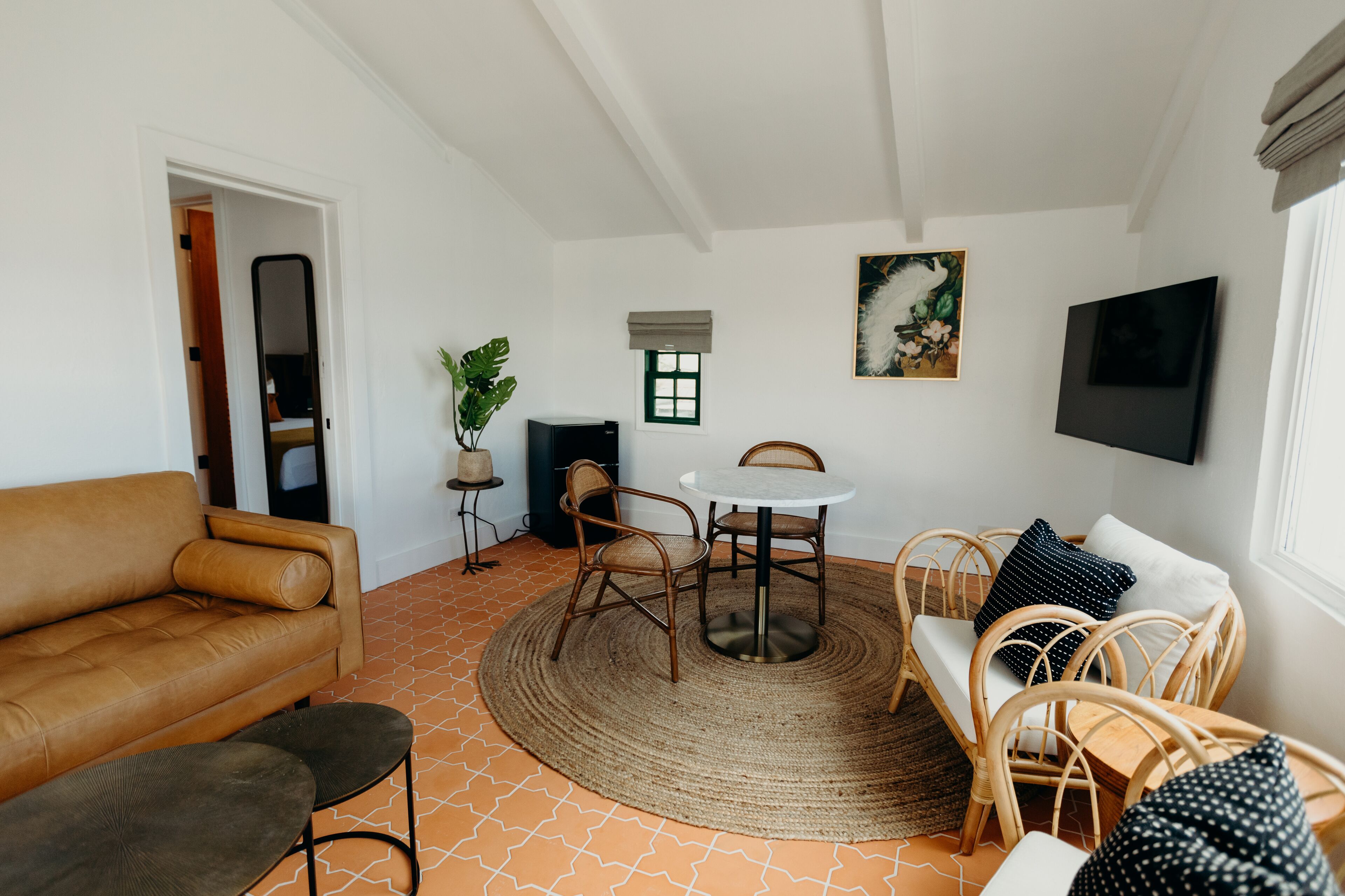 A living room with a brown leather sofa, a round table with chairs, a TV mounted on the wall, and a small window with a view.