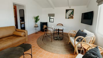 A living room with a brown leather sofa, a round table with chairs, a TV mounted on the wall, and a small window with a view.