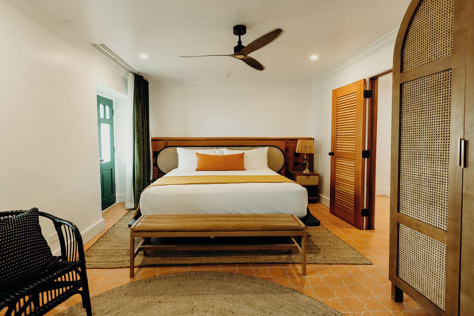 A hotel room with a bed, a ceiling fan, a nightstand, a bench, a chair, and a door with a louvered design.