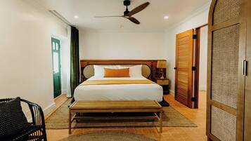 A hotel room with a bed, a ceiling fan, a nightstand, a bench, a chair, and a door with a louvered design.