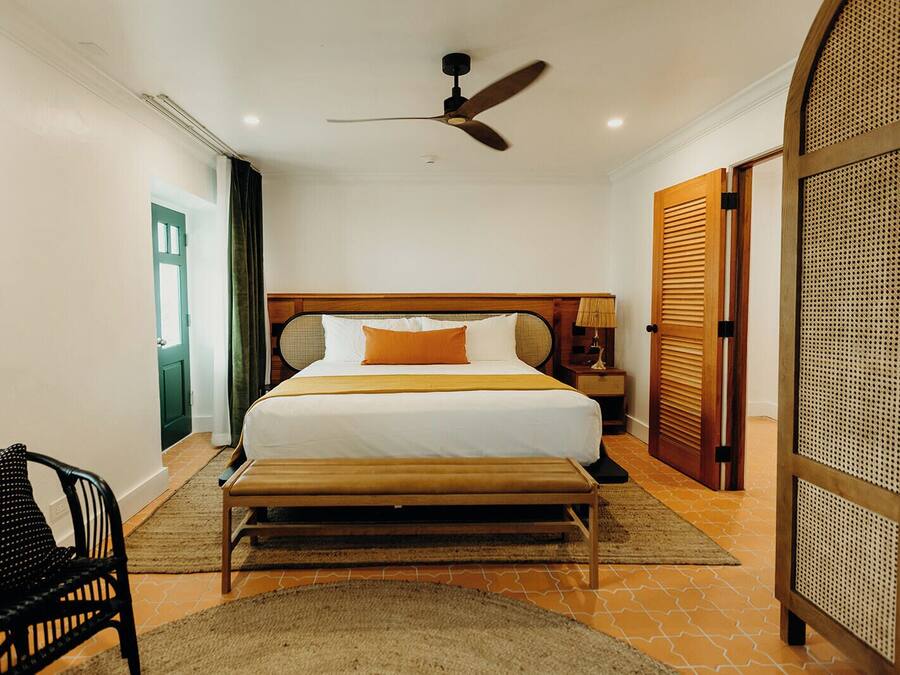 Una habitación de hotel con cama, ventilador de techo, mesita de noche, banco, silla y puerta con diseño persiana.
