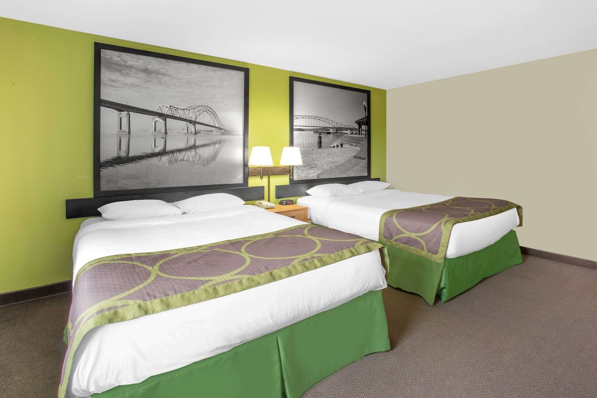 Une chambre d’hôtel avec deux lits, des couvre-lits verts, une table de chevet avec une lampe et deux tableaux encadrés au mur.
