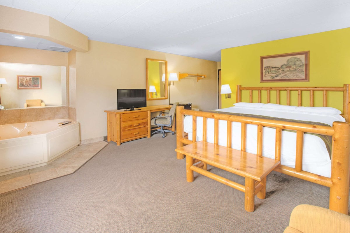 Une chambre d’hôtel avec un lit en bois, un bureau avec un ordinateur, une télévision, une salle de bain avec une baignoire et un tableau au mur.
