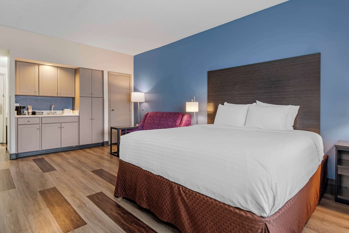 A hotel room with a large bed, a desk, a chair, a kitchenette, and a bathroom.