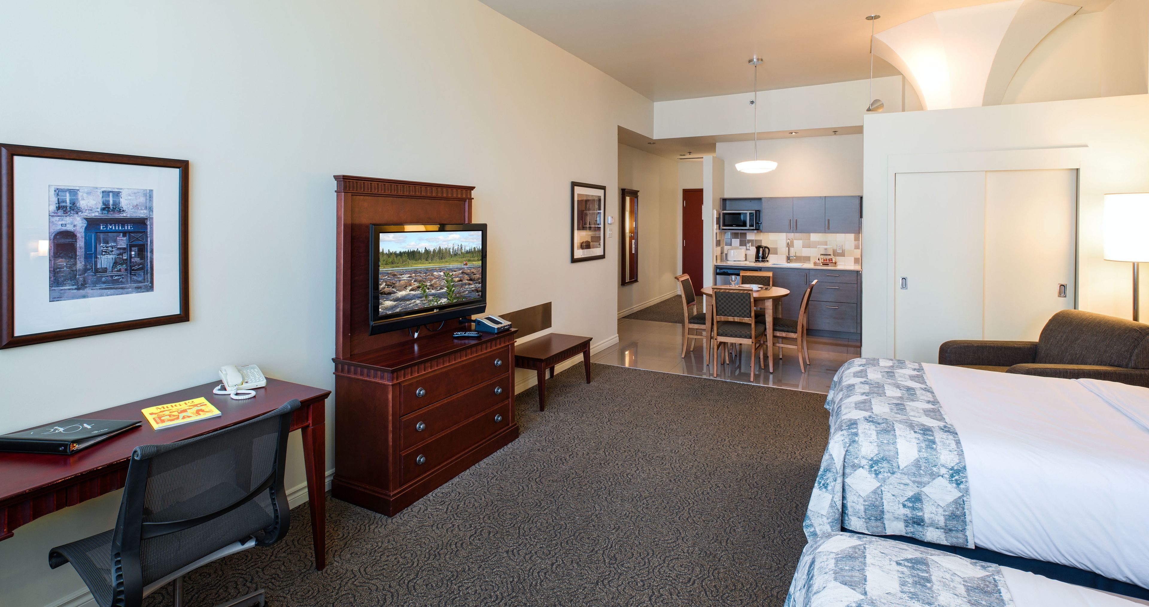 A hotel room with a bed, desk, chair, dresser, television, and a view of the kitchen and dining area.