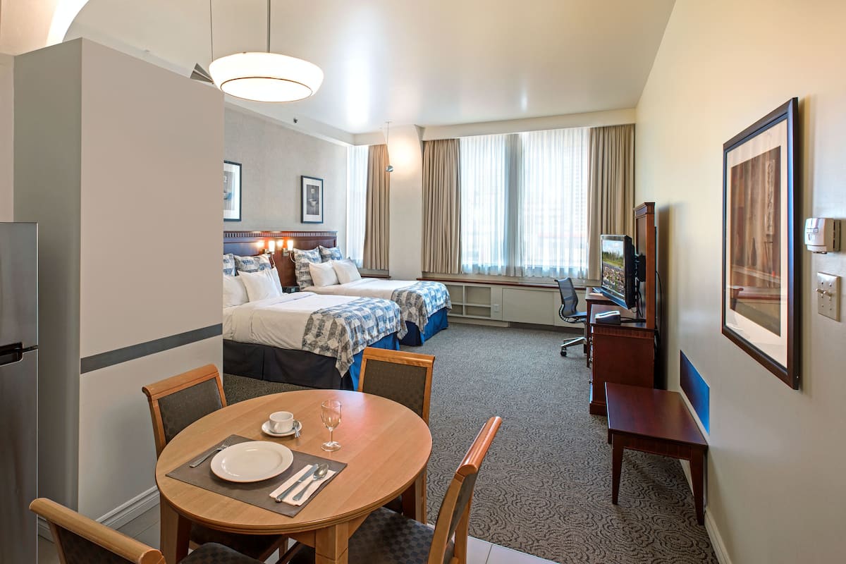 A hotel room with a kitchenette, a dining area, a bed with a headboard, a TV, and a window with curtains.
