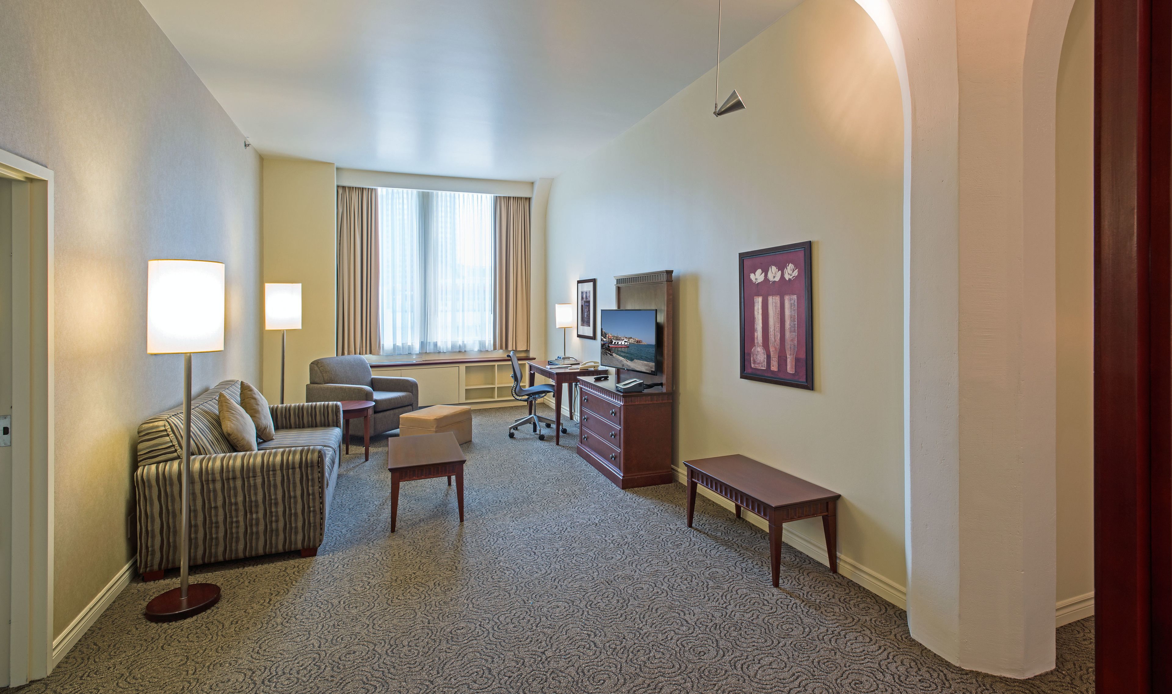 A hotel room with a sofa, armchair, desk, television, and artwork.