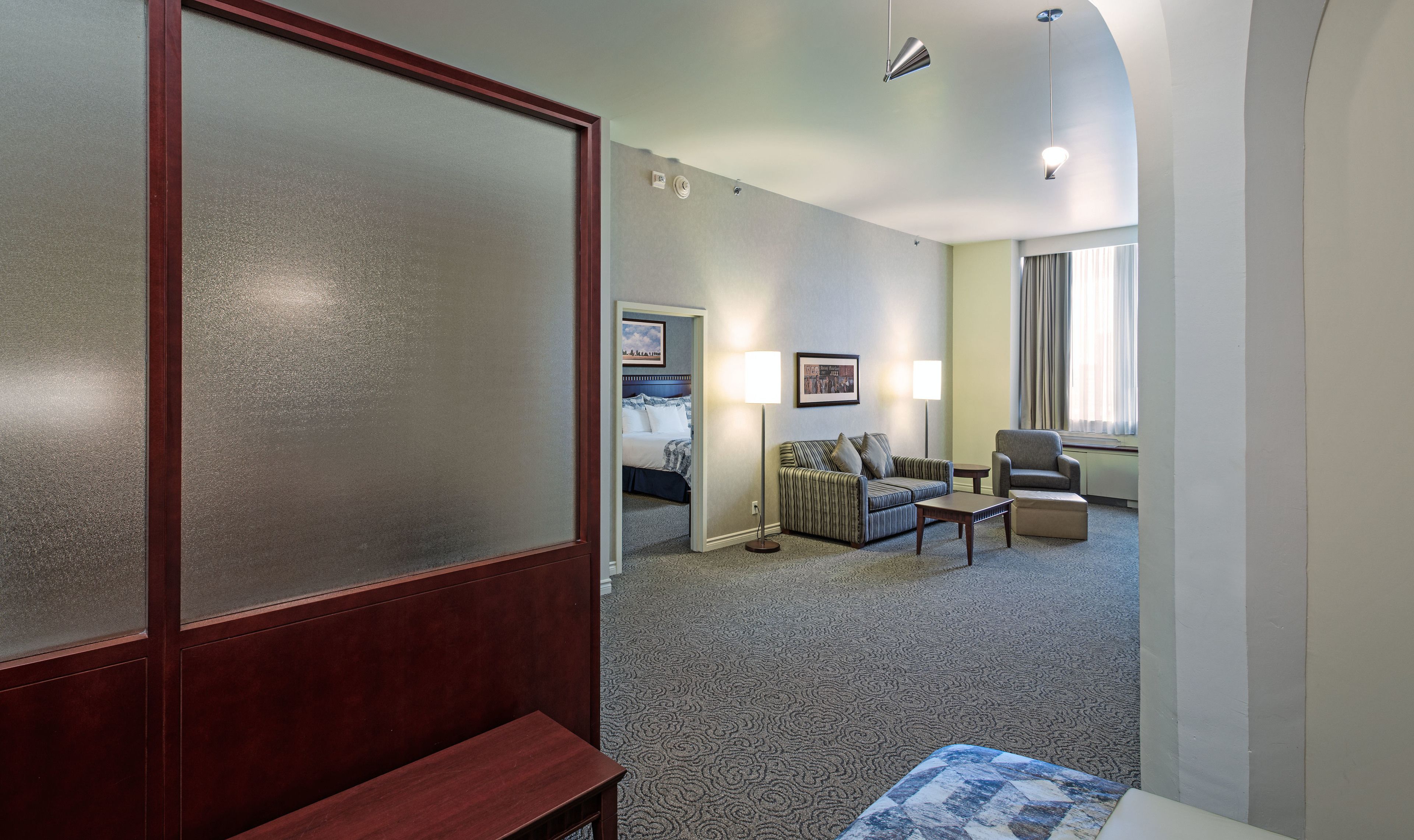 Una habitación de hotel con cama, sofá, sillón, lámpara y ventana con cortinas.