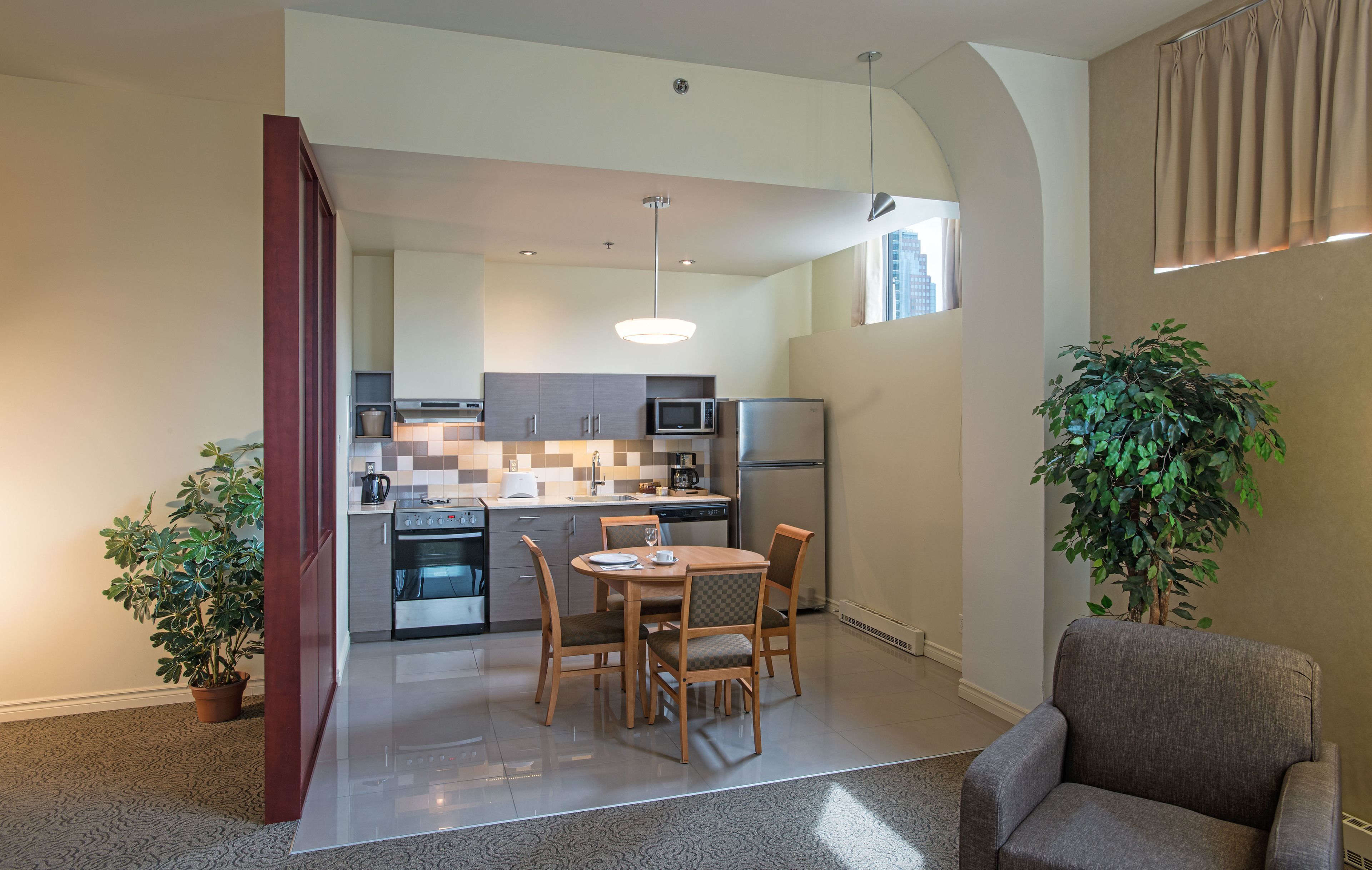 A modern kitchen with a dining area, a large plant, and a grey armchair.