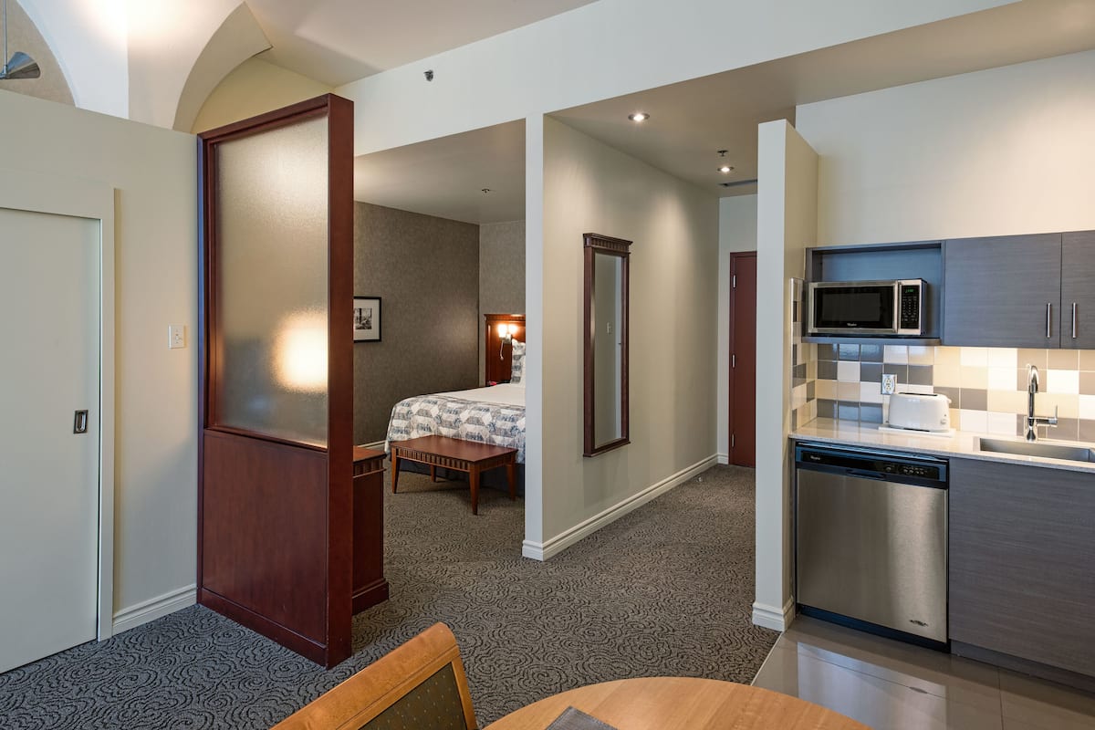 A hotel room with a bed, a desk, a chair, a dining table, a kitchen with a microwave, and a refrigerator.