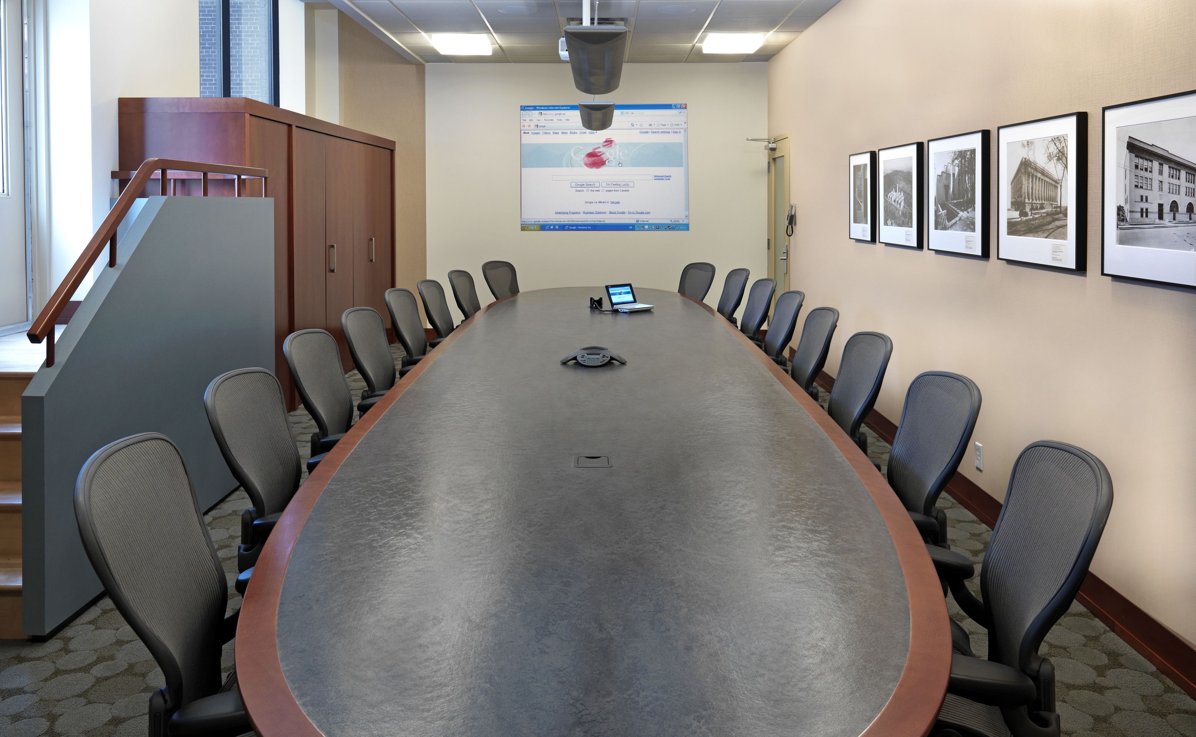 A conference room with a long table, chairs, a laptop, and a mounted screen displaying a webpage.
