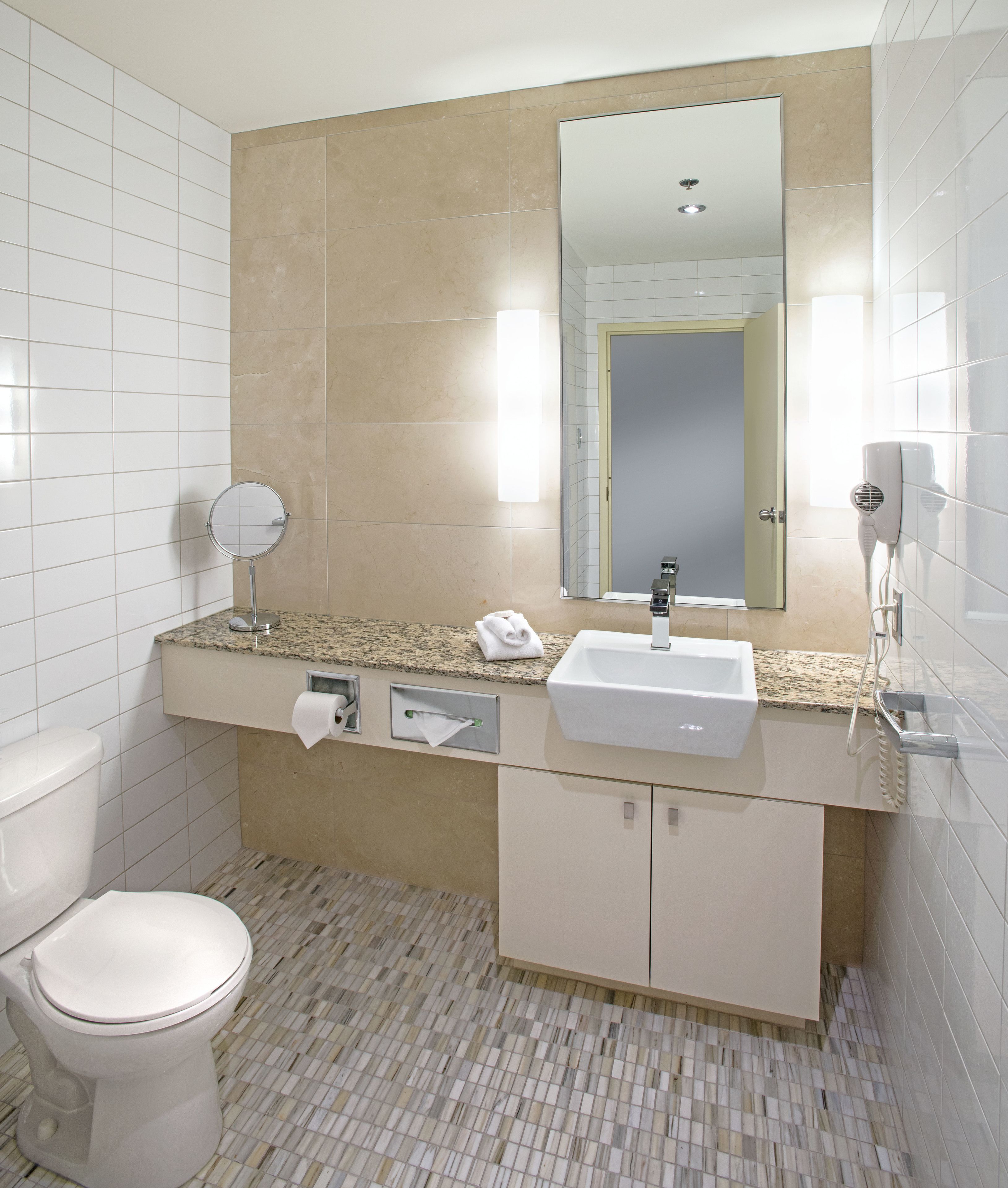 Une salle de bain moderne équipée d’un WC, d’un lavabo, d’un miroir, d’un distributeur de serviettes et d’un sèche-cheveux.