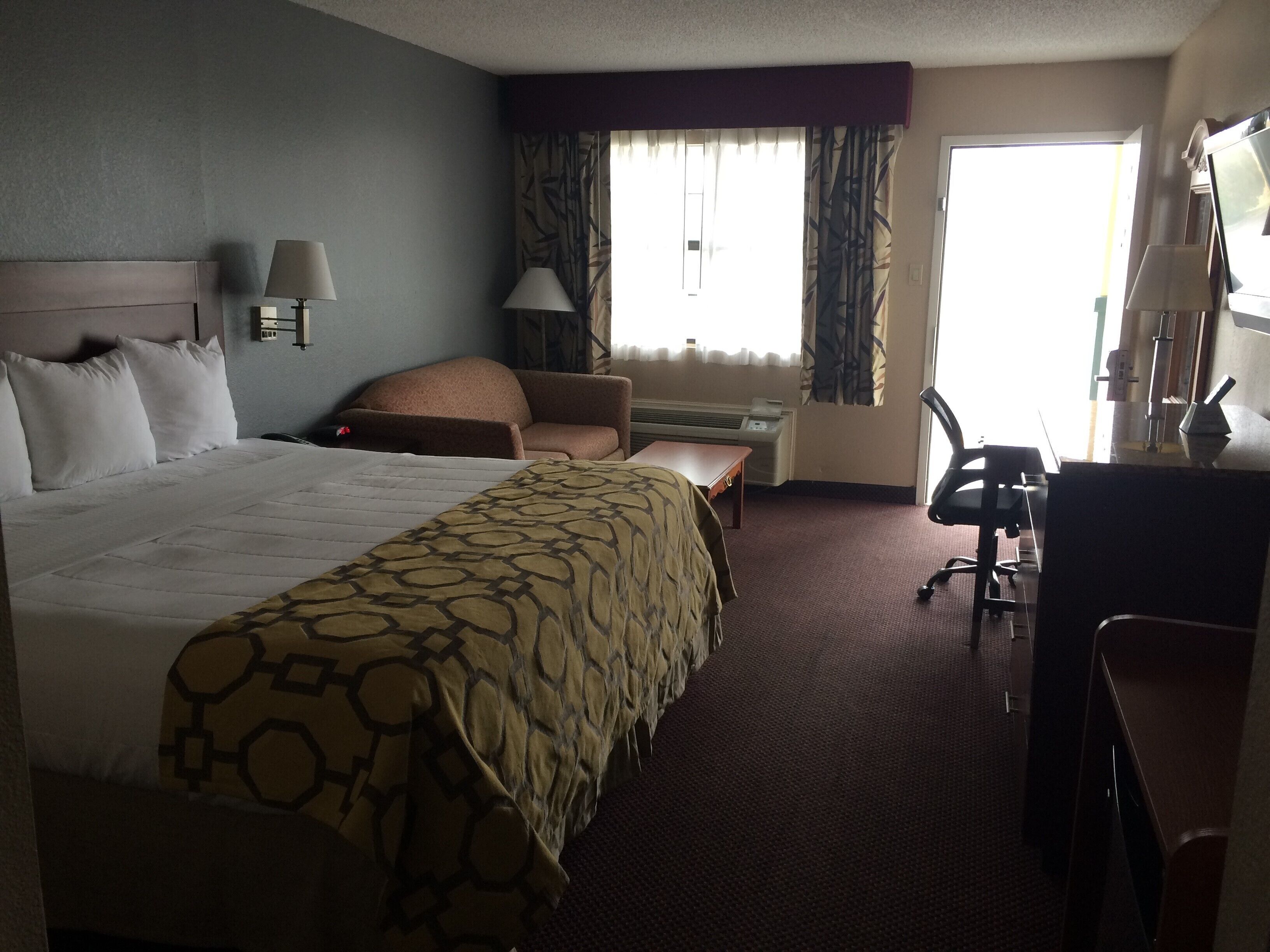 A hotel room with a large bed, a desk, a chair, and a sofa. There is a window with curtains and a wall-mounted air conditioning unit.
