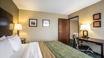 A hotel room with a bed, a desk, two chairs, a door, and framed pictures on the wall.