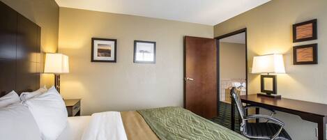 A hotel room with a bed, a desk, two chairs, a door, and framed pictures on the wall.