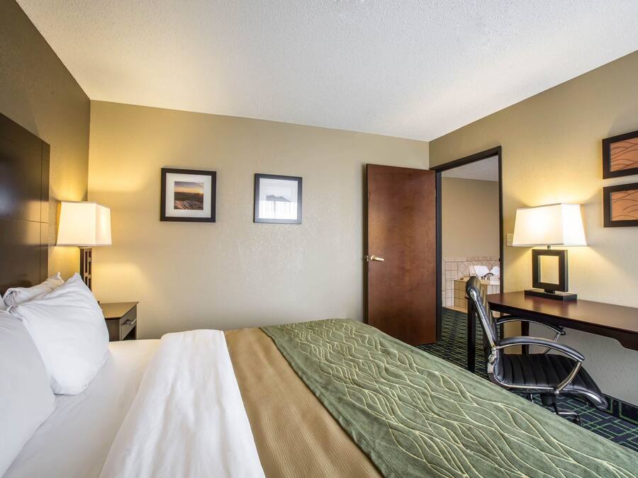 A hotel room with a bed, a desk, two chairs, a door, and framed pictures on the wall.