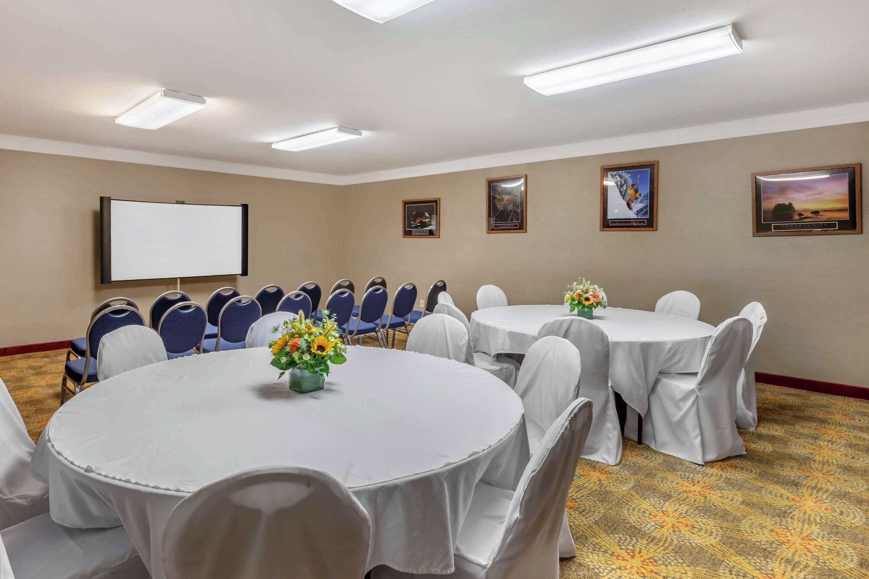 Une salle de conférence équipée de tables rondes, de chaises, d’un écran de projection et de tableaux encadrés aux murs.