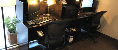 Un cubículo de oficina moderno con dos escritorios, cada uno equipado con una computadora, teclado y ratón. Hay una lámpara en un escritorio y una pequeña planta en maceta en el suelo.