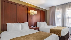 A hotel room with two beds, a wooden headboard, a nightstand with a lamp, and a window with curtains.