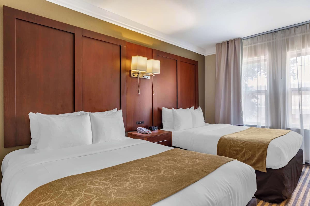 Ein Hotelzimmer mit zwei Betten, einem hölzernen Kopfteil, einem Nachttisch mit Lampe und einem Fenster mit Vorhängen.