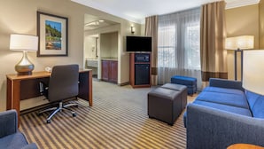 A hotel room with a checkered carpet, a desk with a lamp, a television, a sofa, and a small kitchenette in the background.