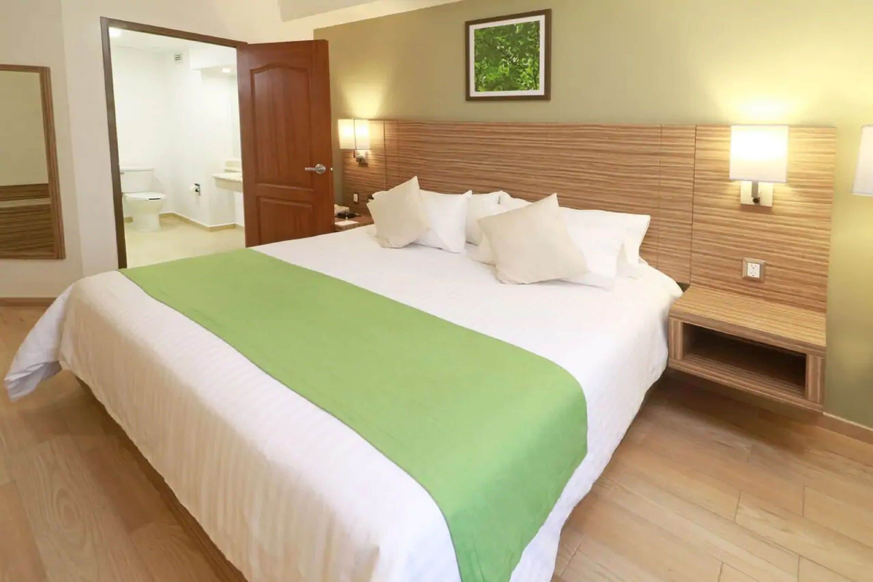 Habitación de hotel con una cama grande, un plaid verde, dos lámparas de noche, un cabecero de madera y un espejo en la pared.