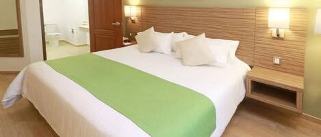 Habitación de hotel con una cama grande, un plaid verde, dos lámparas de noche, un cabecero de madera y un espejo en la pared.