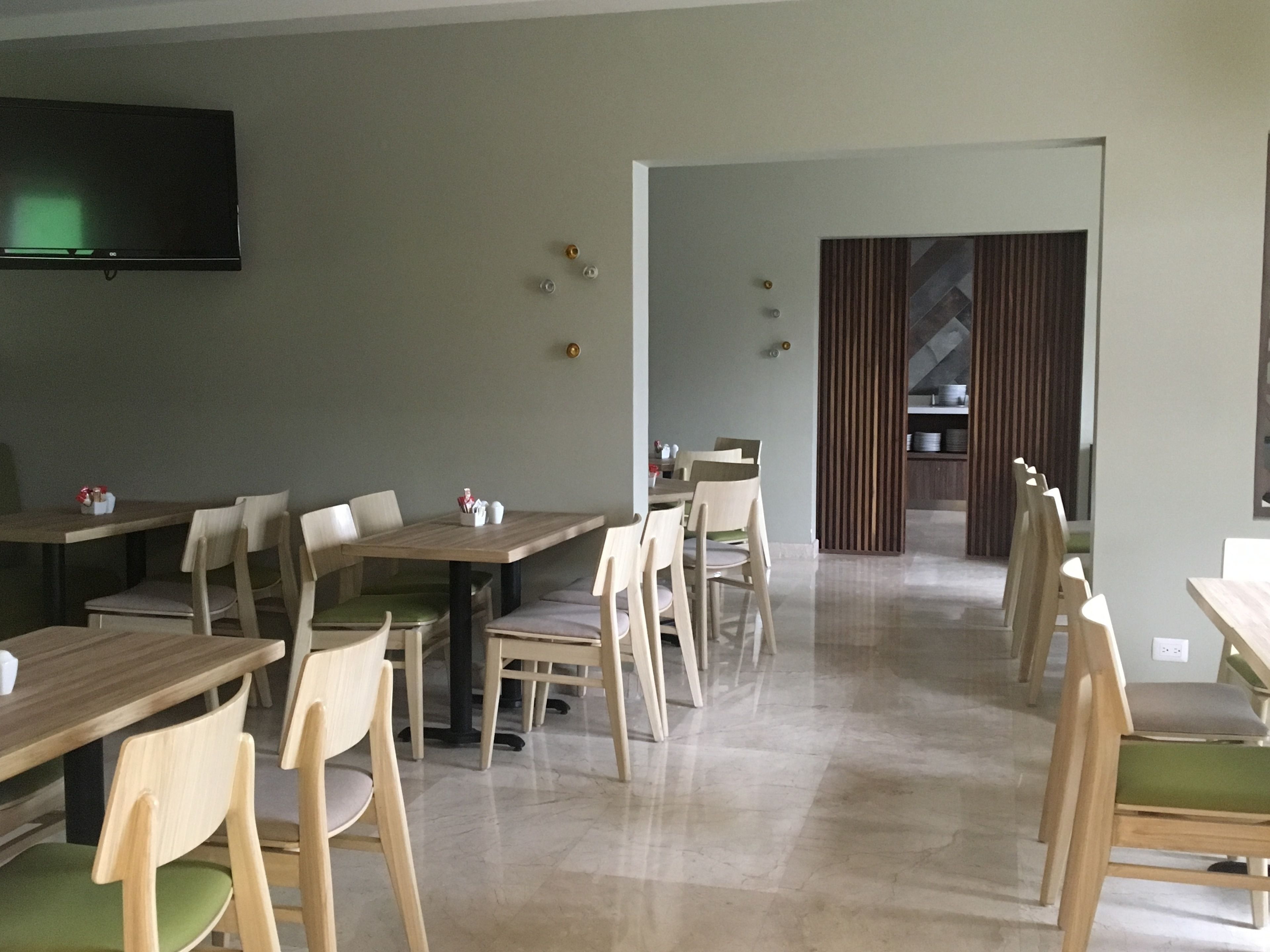 A spacious dining area with multiple wooden tables and chairs, a flat-screen TV mounted on the wall, and a modern kitchen visible through a glass partition.