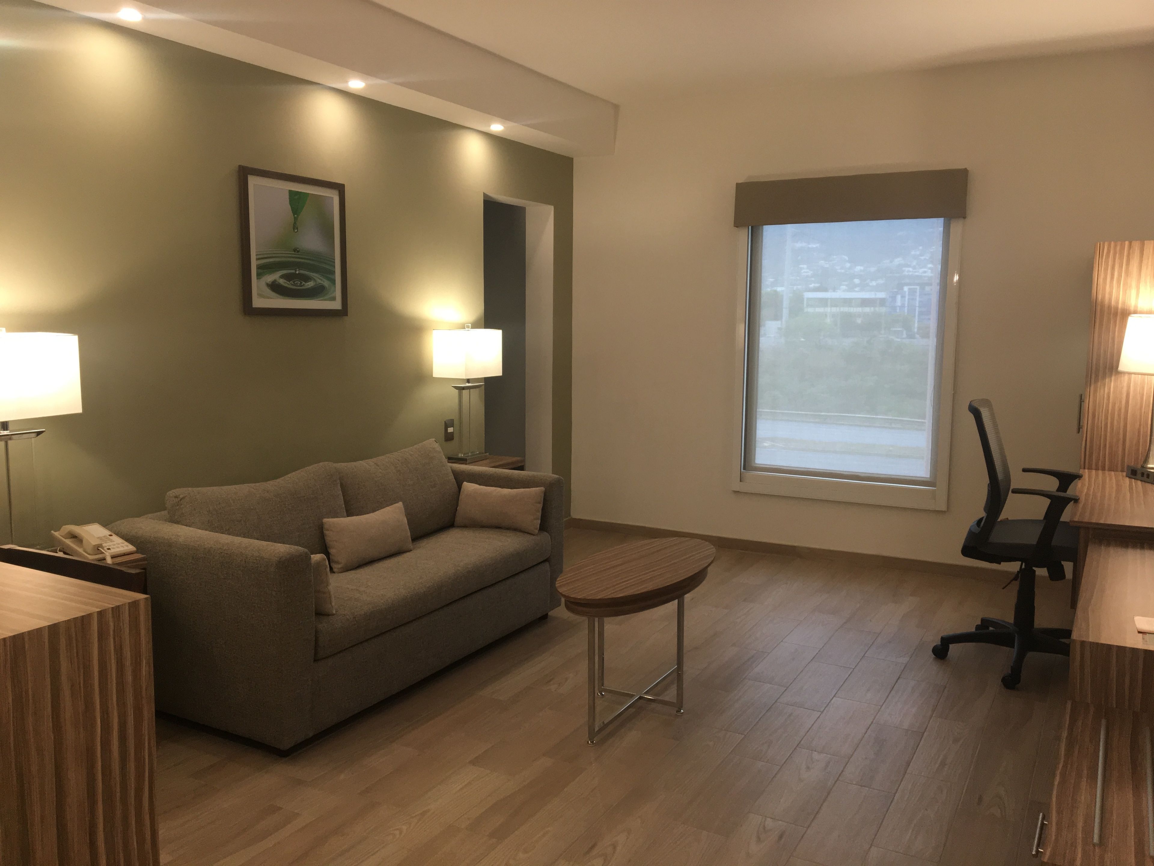 A modern hotel room with a sofa, a small round table, a desk with a chair, a lamp, and a large window with a view.