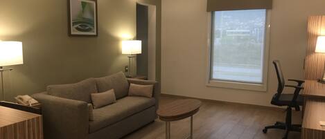 A modern hotel room with a sofa, a small round table, a desk with a chair, a lamp, and a large window with a view.