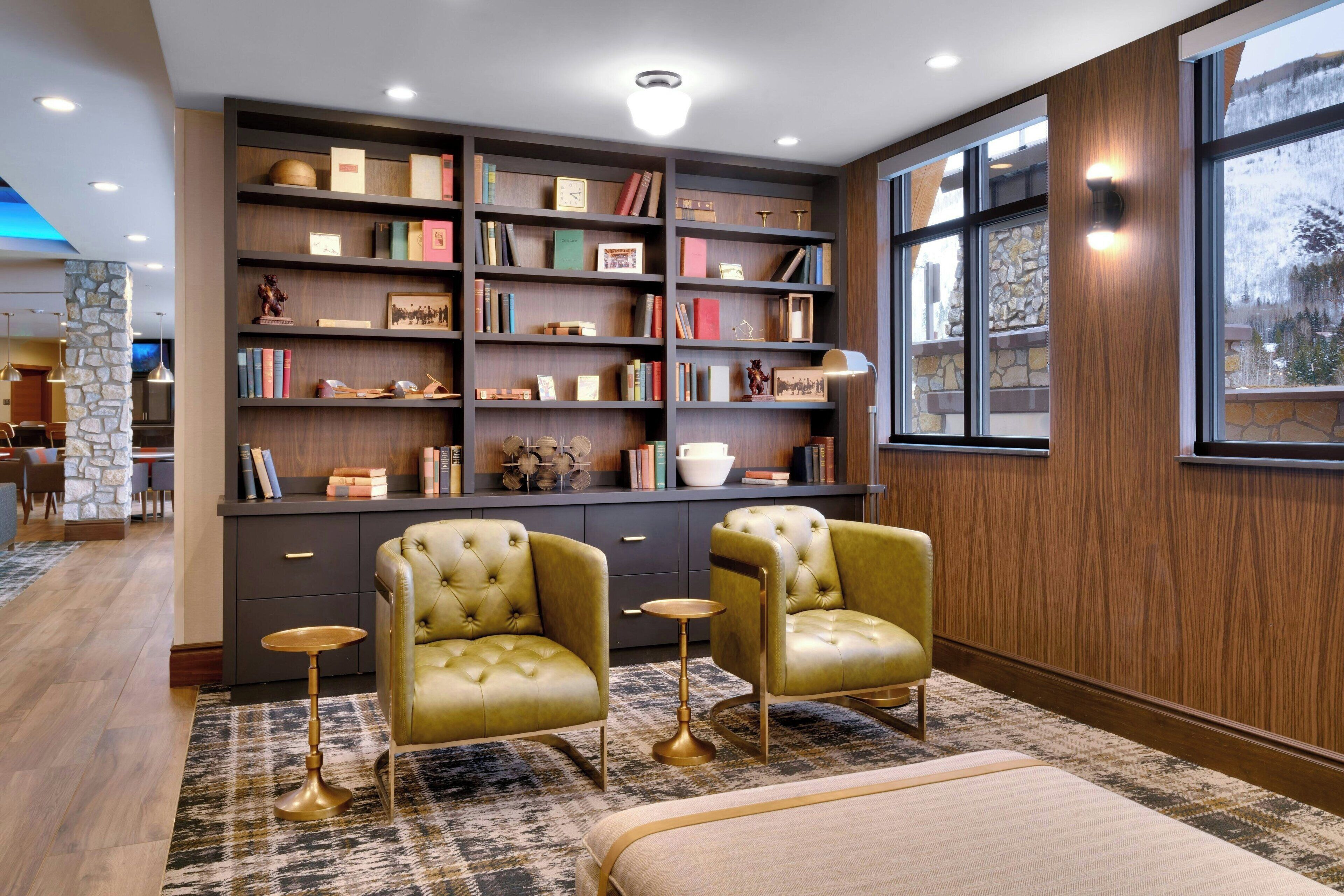 A modern library with bookshelves, two leather armchairs, a round table, and a large window with a view.