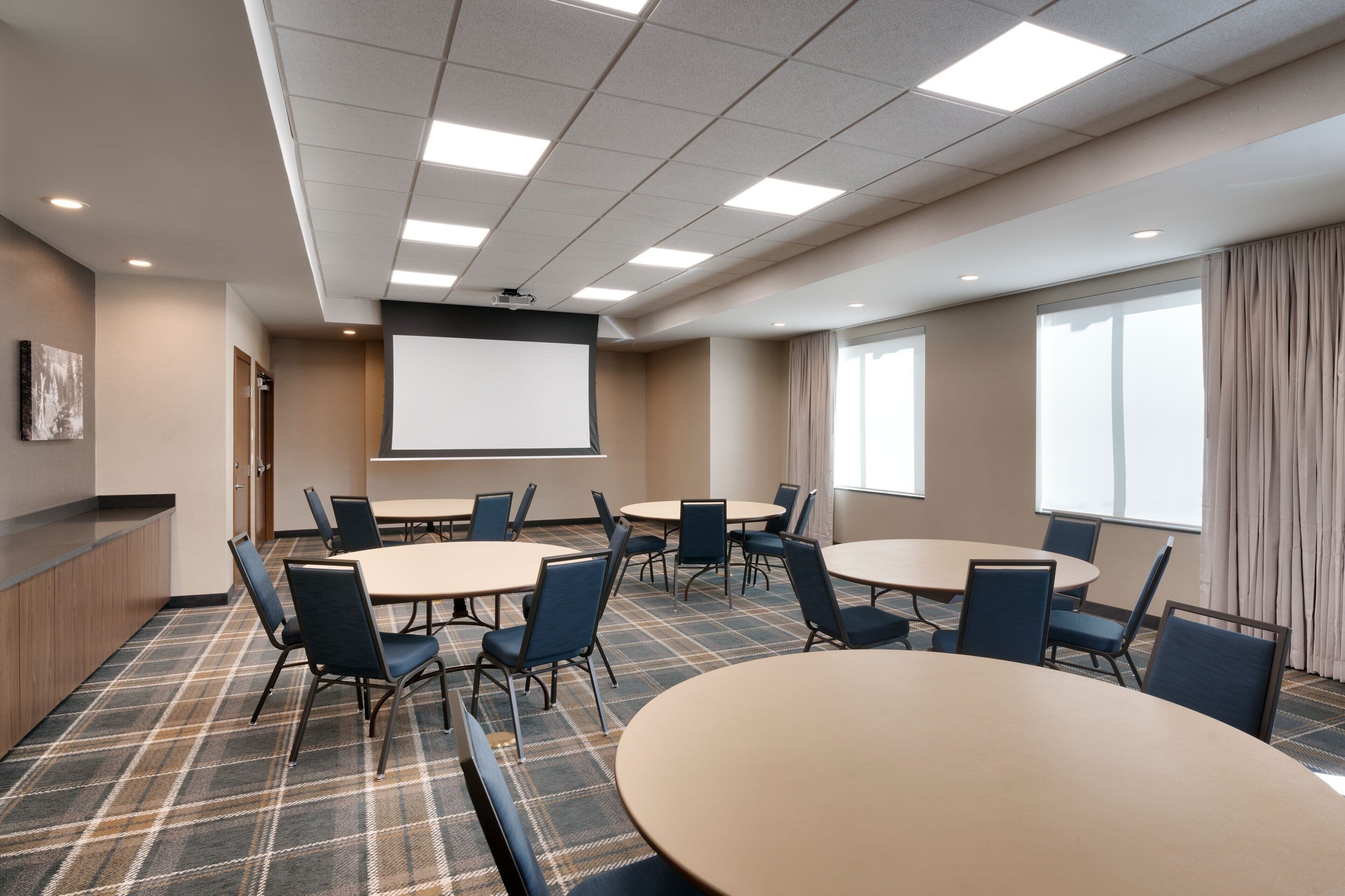 A conference room with multiple tables and chairs, a projector screen, and a large window with curtains.