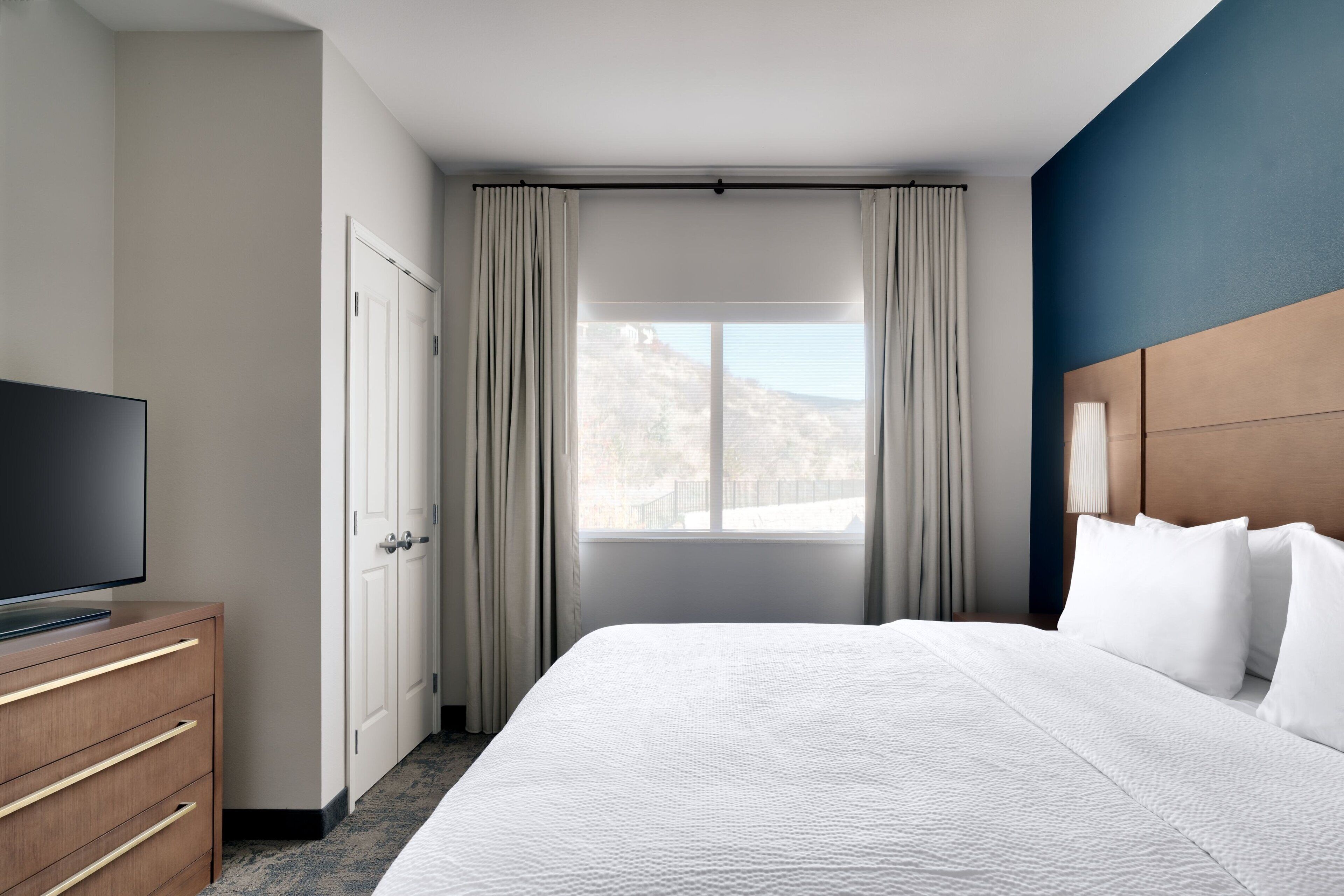 A hotel room with a bed, a TV on a wooden dresser, a window with curtains, and a view of a mountain.