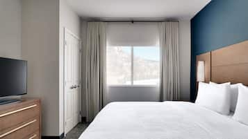A hotel room with a bed, a TV on a wooden dresser, a window with curtains, and a view of a mountain.