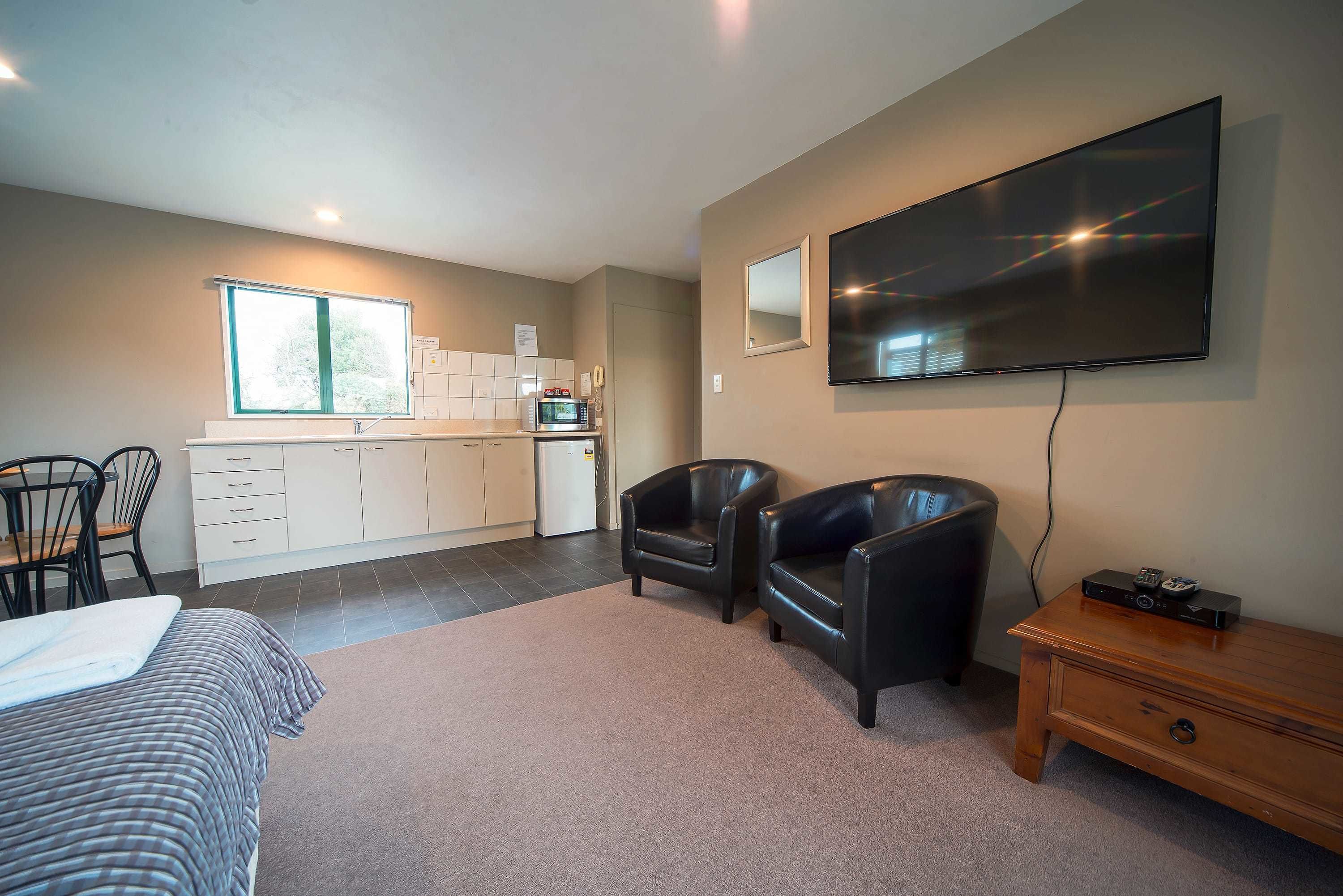 A modern hotel room with a bed, a desk, a flat-screen TV, two leather chairs, a wooden nightstand, and a kitchenette.