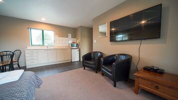 A modern hotel room with a bed, a desk, a flat-screen TV, two leather chairs, a wooden nightstand, and a kitchenette.